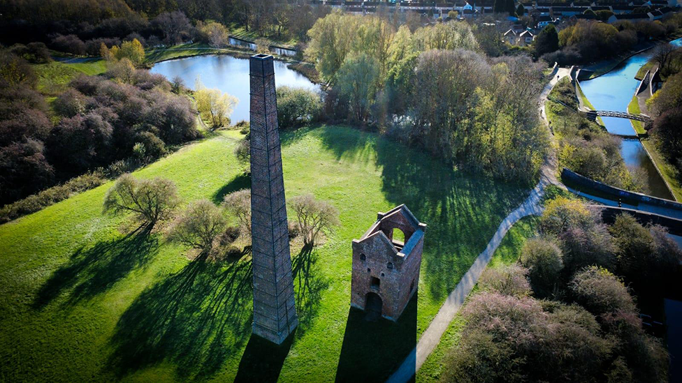 An aerial view of a tall chimney and derelict building in a field in Sandwell. Behind the chimney is a large pool