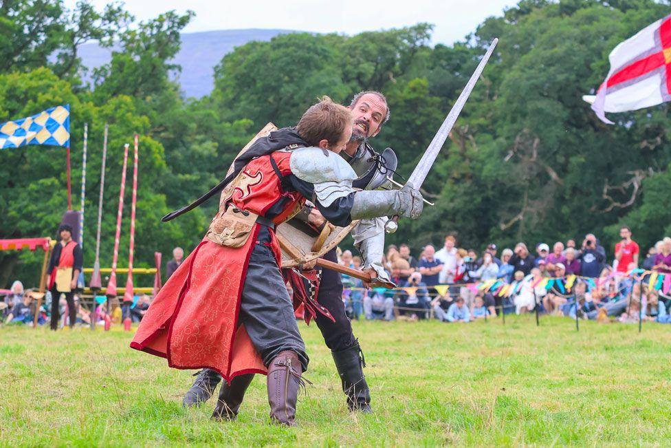 Two men in historic costume stage a sword battle in front of large crowds