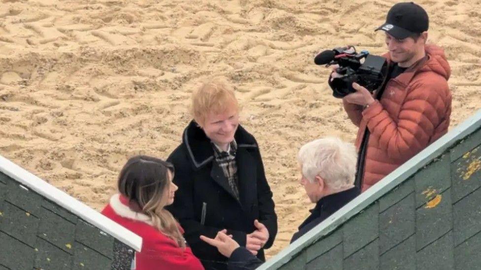 Claudia Jessie can be seen from behind she has brown long hair and has a red jacket on with a white fur trim. She is stood next to Ed Sheeran who is smiling and is wearing a black coat. Alongside on the beach is Richard Curtis who has his back to the camera and is wearing a black coat and a man with an orange coat and black cap who is holding a camera. 