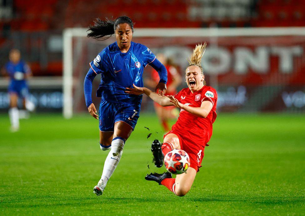 A woman with dark long hair, wearing a blue football kit with white socks runs towards a ball and a woman with fair hair wearing a red football kit lunges with her right foot towards the ball. The woman in red is shouting as she falls towards the grass pitch. 