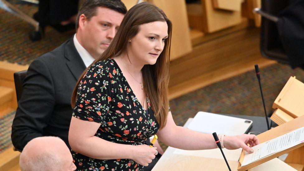 Natalie Don-Innes, a woman with long brown hair, speaks at a podium in the Scottish Parliament chamber. She is wearing a black dress decorated with a floral pattern. 