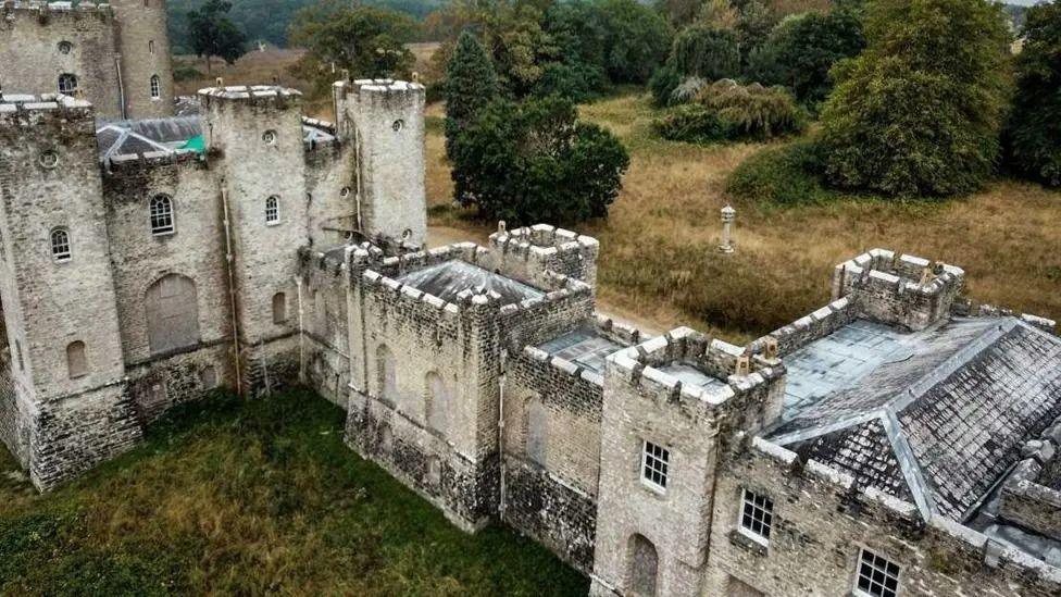 An aerial view of a castle with white stone walls that are stained grey, with turrets and towers rising over the main body. It's in a rural location with trees and grass on both sides.