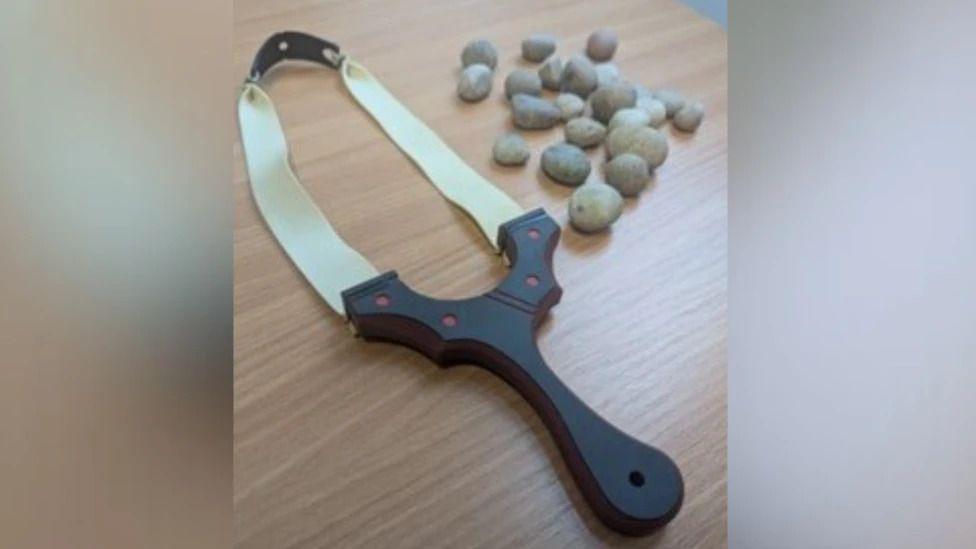 An image of a catapult sent over by Kent Police. The catapult has a black handle and white elastic. A number of stones are pictured to the right of the catapult.