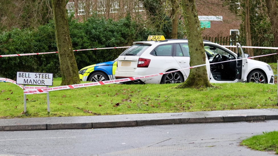 A white estate car with a taxi sign sits in a car park. There is a police cordon tied to a street sign for "Bell Steel Manor". A marked police car is parked behind the taxi. There are a variety of trees and patches of grass at the perimeter.
