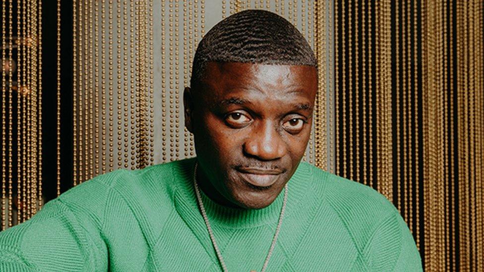 Akon wearing a green top with his hands joined together at the fingertrips. He is wearing a gold necklace as he looks into the camera. The background is shiny gold pearls dropping from top to bottom, as his right elbow is perched on a table.