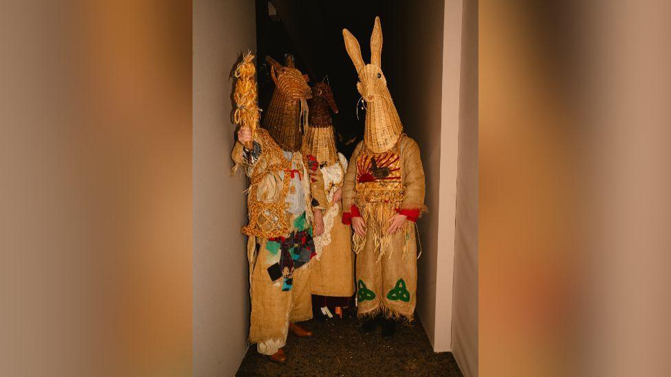 A portrait photo featuring three people dressed up in the traditional mumming outfits. All three are wearing woven headpieces depicting animals, and are wearing clothing made out of various textures such as hessian and repurposed potato sacks. They are embellished with various celtic symbols, embroidery and patchwork.