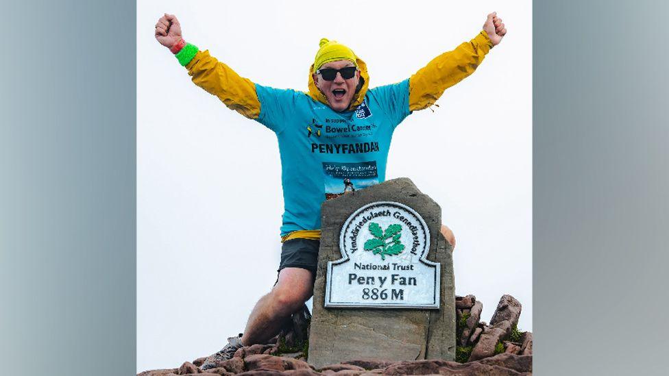 A man at the top of Pen y Fan. The stone at the top shows it is Pen y Fan and is 886 metres. There are rocks around the main headstone. The man has a blue t-shirt and under a yellow long sleeved shirt with a yellow hat, black sunglasses and black shorts. His arms are outstretched like his is cheering. 