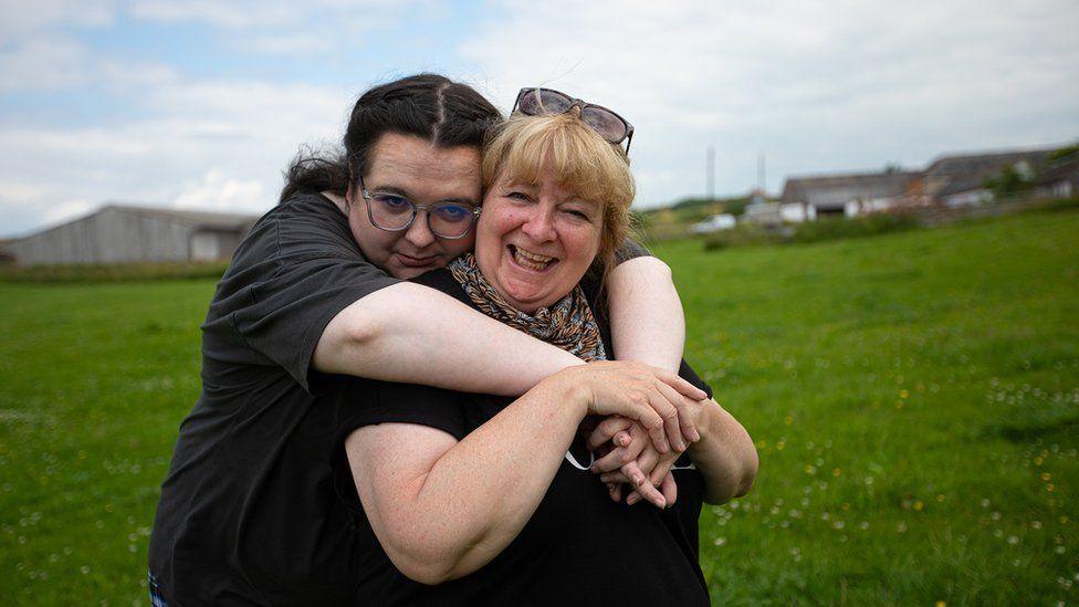 Ashley Storrie and Janey Godley standing in a field. Ashley has har arms around her mother, standing behind her