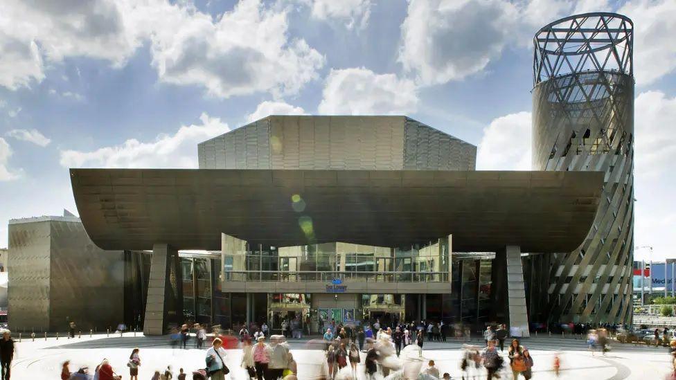 The Lowry arts centre, a steel and glass-clad building facing a paved public plaza. Crowds can be seen milling around the building. 