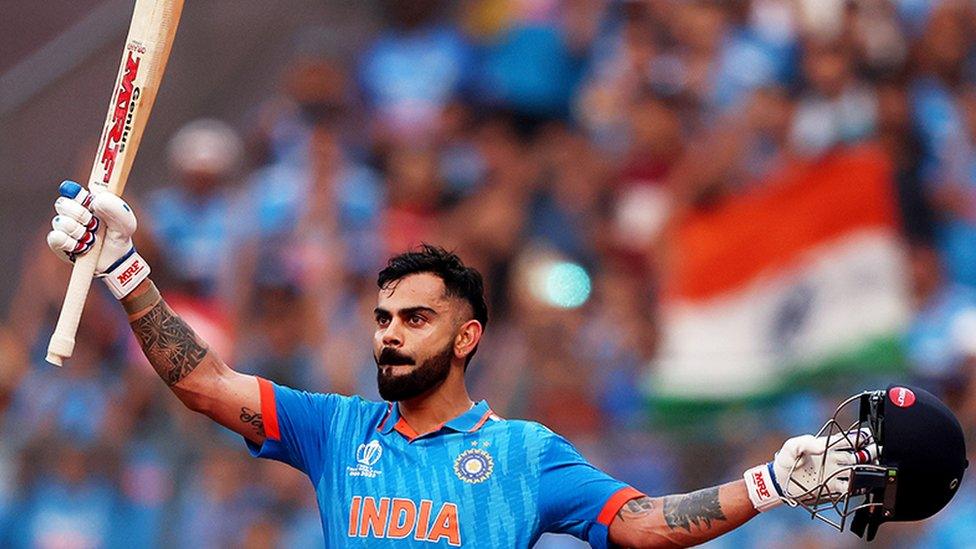 Virat Kohli, an Asian man, holding a cricket bat in his right hand, raised, with his black helmet in his left hand. He is celebrating a milsetone, wearing a blue India jersey with "INDIA" printed in orange. He is wearing white cricket gloves. The background is blurred with the orange, white and green India flag visible.