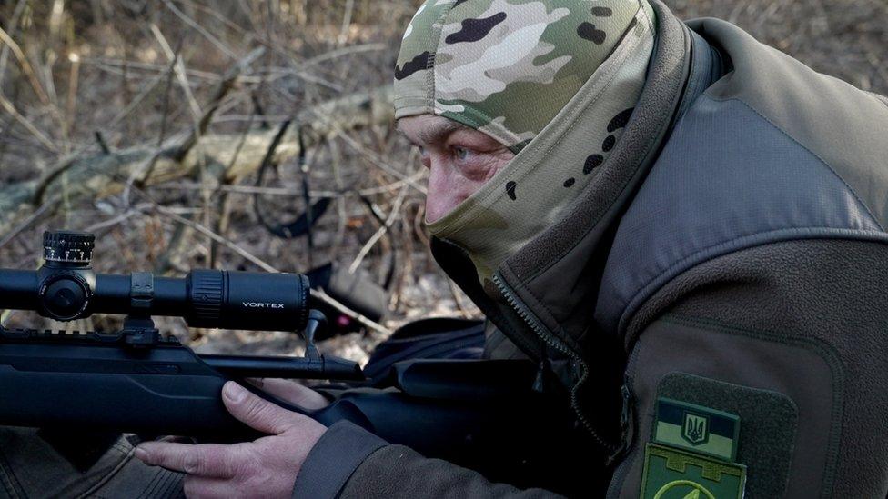 Alex, wearing a balaclava and hood, practices with his sniper rifle