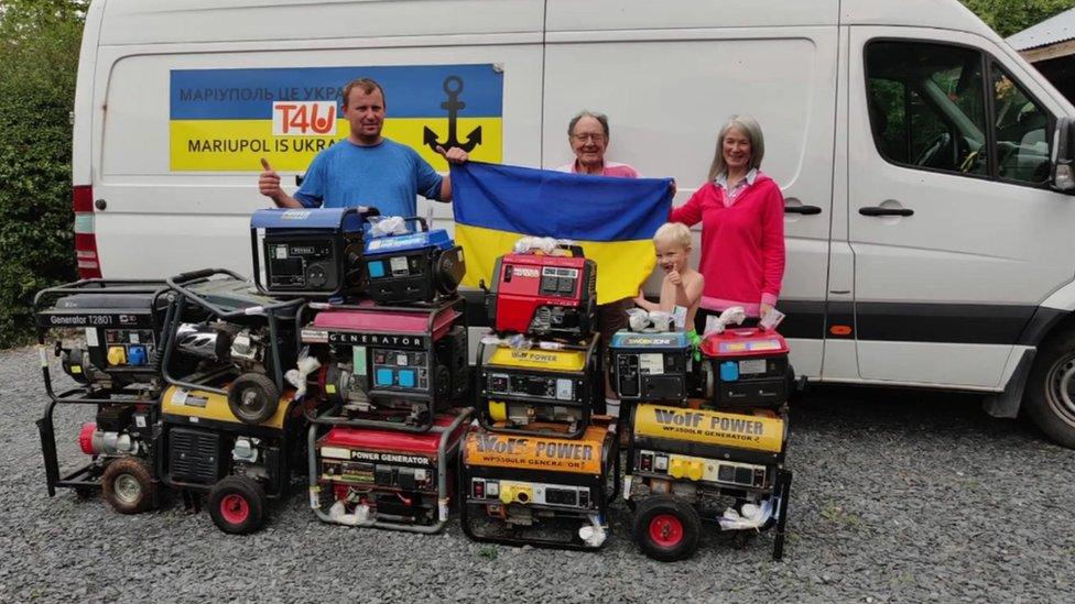 Generators stacked outside van with people holding Ukrainian flag