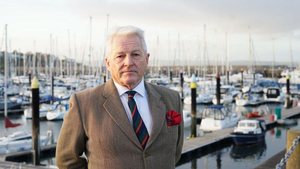 Col Tim Collins, wearing a suit, looks into the camera on Bangor marina with boats behind him