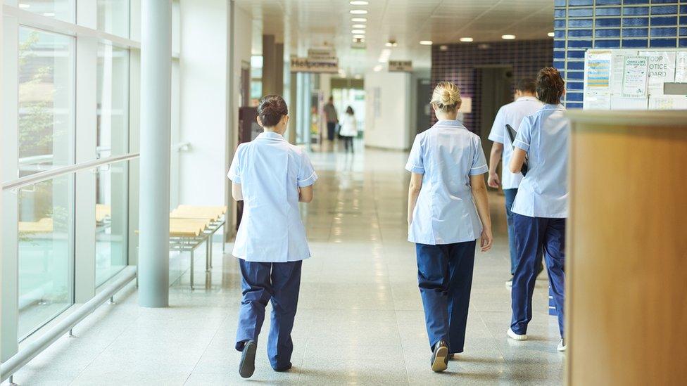 A group of four young trainee nurses including male and female nurses, walk away from camera down a hospital corridor. They are wearing UK nurse uniforms of trousers and tunics.