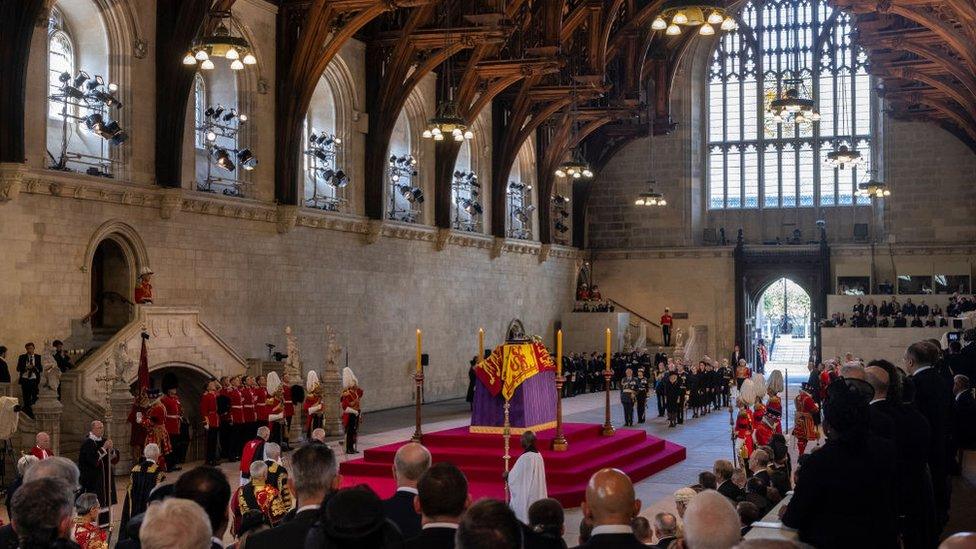 the queen's coffin lying in state in westminster hall surrounded by guards with members of the royal family looking on