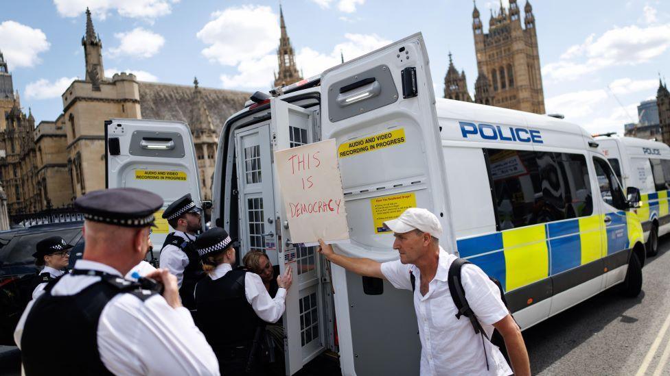 Four police officers stand at the back of an open police van where a person appears to be being put in the back. Next to them a man holds a sign saying 'This is democracy'. In the background the Palace of Westminster can be seen.