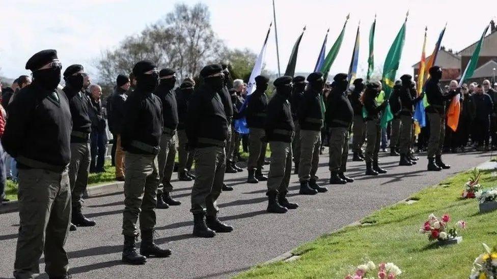 A number of masked men wearing berets , combat trousers and black jumpers stand at a commemoration holding Irish nationalist flags