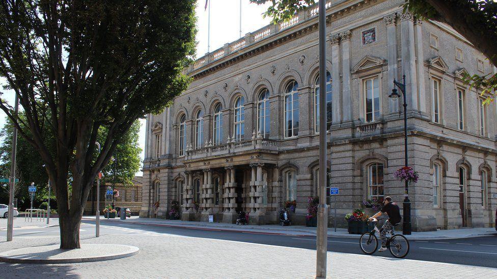 An old beige building with several oval windows across a street. A cyclist is on the road but there are no other vehicles or people.