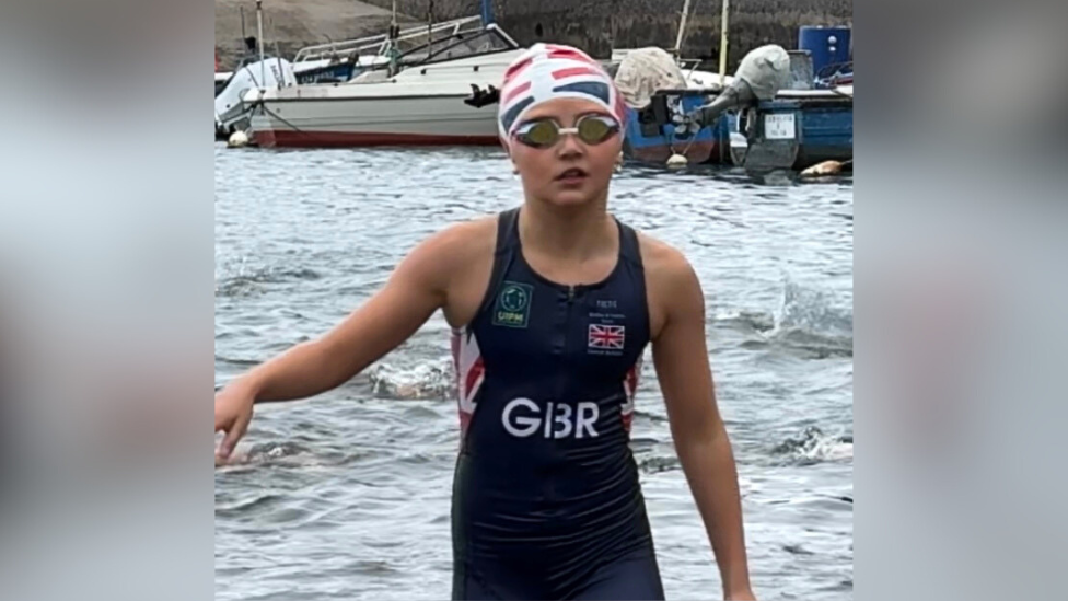 Evie in a swimming cap and goggles coming out of the water 