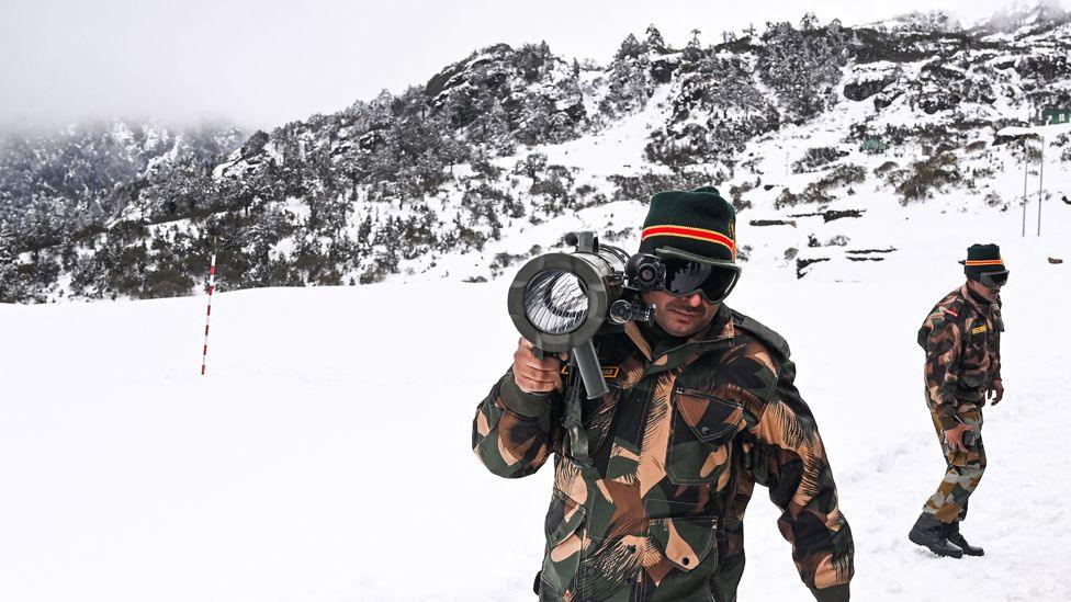 Two Indian soldiers in camoflage in the snow in the Himalayan mountains. They are wearing goggles and one is carrying a shoulder mounted rocket launcher