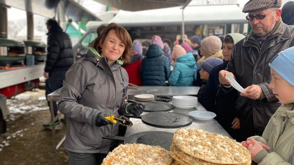 A woman serves pizza to people lined up at a counter. She has brown hair and is wearing a grey jacket and dark trousers. She is holding a pizza cutter. Several pizzas are on the counter in the front of the photograph.