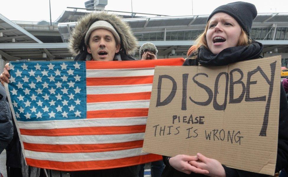 Demonstrators with a US flag and a sign urging protest stand at JFK airport