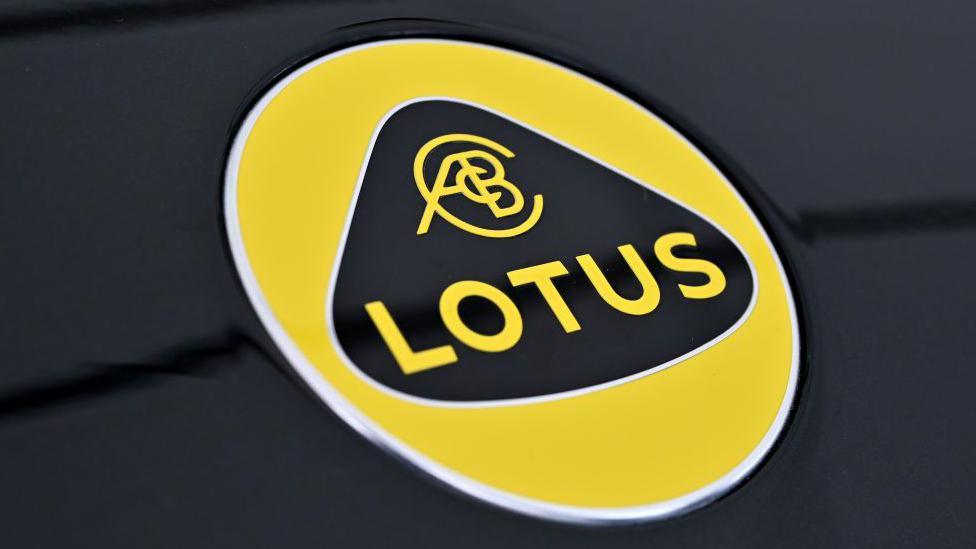 The Lotus logo. It has yellow writing on a black background surrounded by a triangular, curved shape. That is then surrounded by a yellow circle. The logo sits on a black, metallic surface.