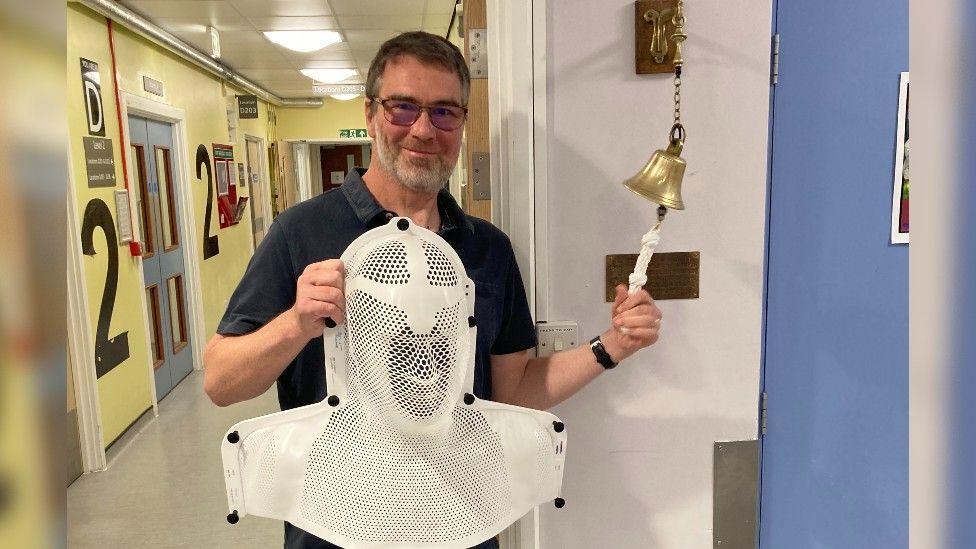 Guy Walker ringing the bell after getting the all clear from his cancer treatment. He is holding the surgical mask he wore in recovery from the operation and wearing glasses and a short sleeve navy top. The hospital corridor behind him has yellow walls and level 2 signs.