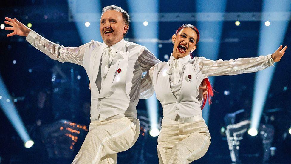 Chris McCausland and Dianne Buswell dancing on Strictly in matching white  suits, smiling and arm in arm, each with their other arm outstretched and with a leg raised.