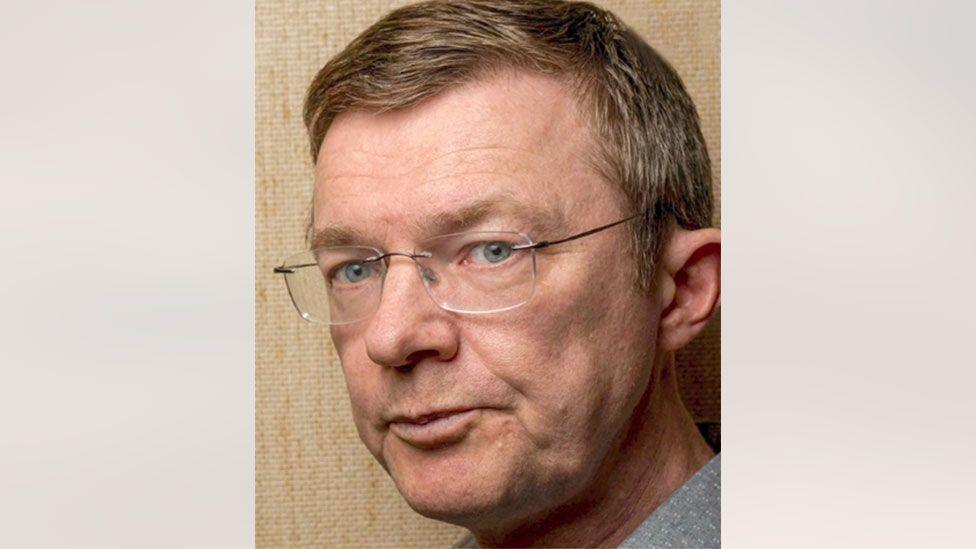 Arne Bye seen on a beige background. He has light hair and blue eyes, and wears glasses.
