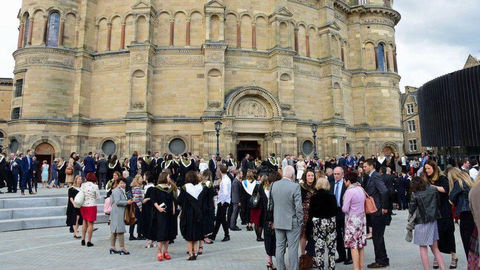 A yellow sandstone building with graduands standing outside in robes with members of their families.