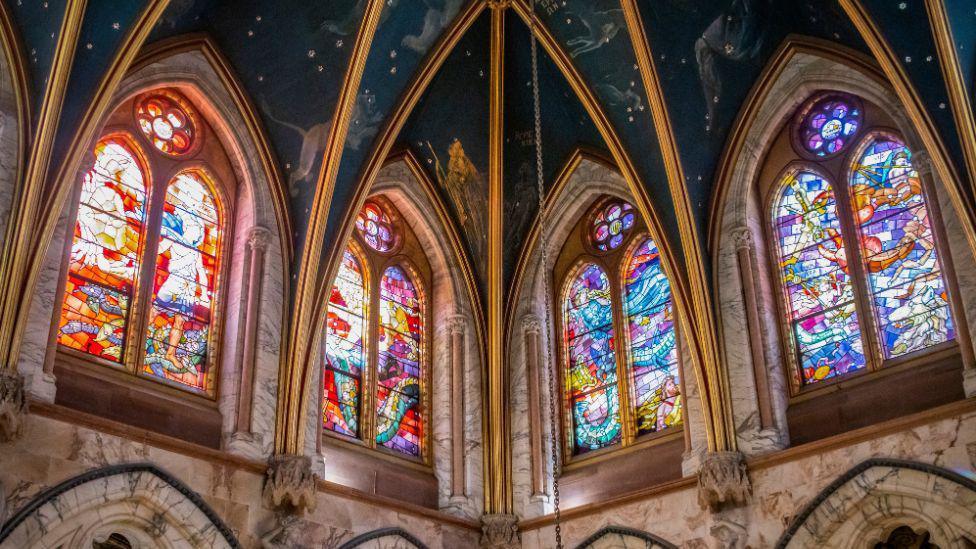 The interior of a Gothic-style church featuring tall, pointed arches and colourful stained-glass windows that fill the space with vibrant light and intricate patterns.