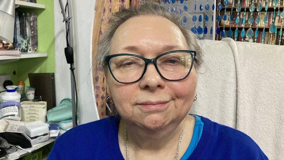 Janet Warner-Waite looks at the camera while sitting in a chair wearing glasses and a blue jumper