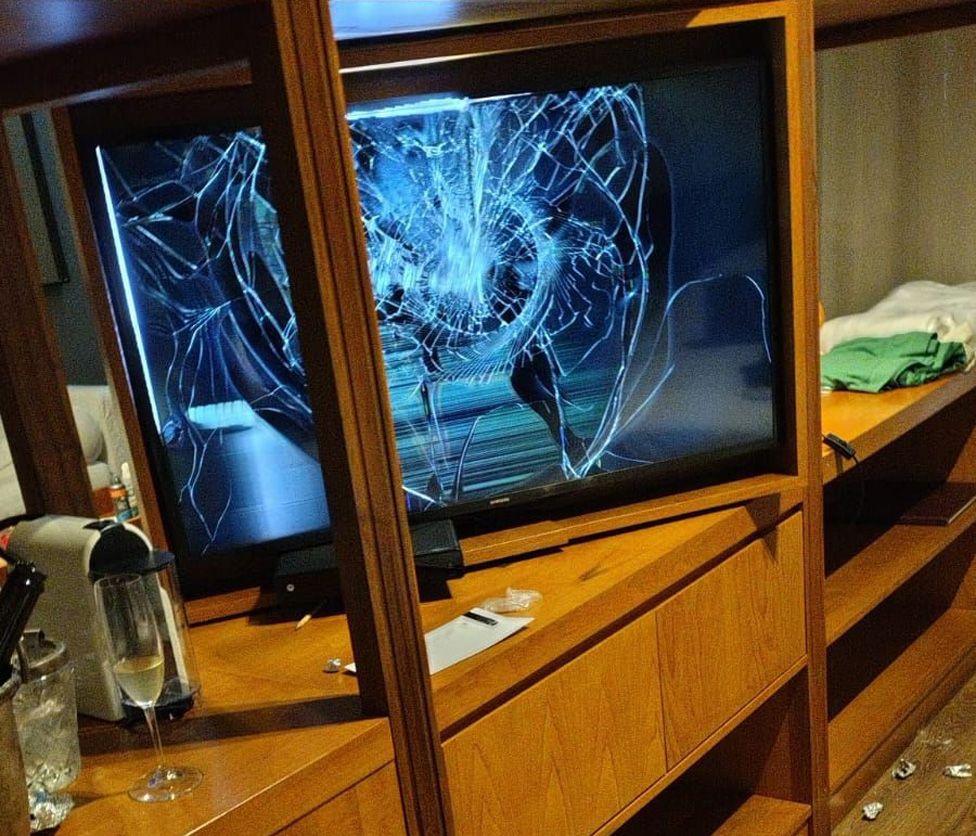 A TV set with its screen smashed, on a table with a glass of champagne and other items strewn around 