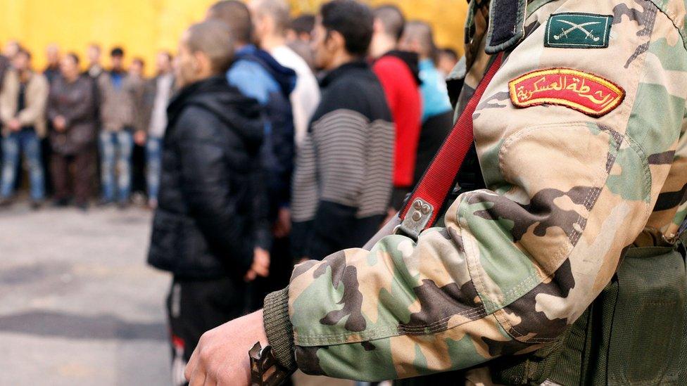 Syrian military police officer stands next to men preparing to being their military service after fleeing rebel-held Aleppo (11 December 2016)