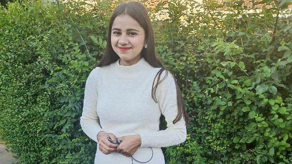 Harshita Brella smiles at the camera as she is photographed outside. There is a large green hedge behind her. She is wearing a beige polo-necked jumper and has long dark hair. She is wearing earrings.