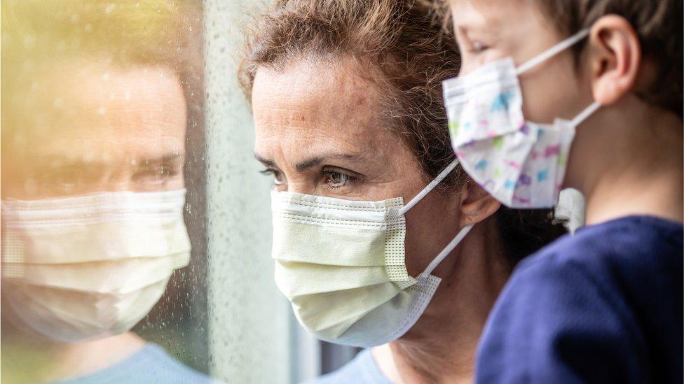 A parent wearing a face mask looks out of a window while holding a child also wearing a mask