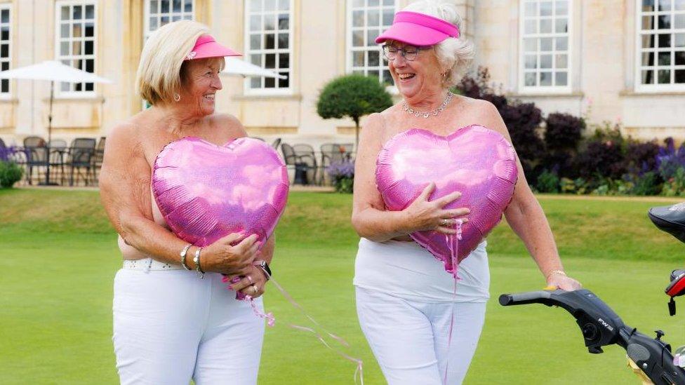 Two women wearing golf caps and white trousers hold large pink hearts in front of their upper bodies.