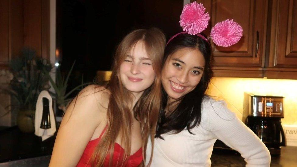 Girlhood founders Sophia Rundle and Mia Sugimoto. Sophia, on the left, has long dark blonde hair which she wears loose. Her eyes are closed as she smiles at the camera and she wears a strappy red dress and leans into Mia, who stands beside her. Mia has long dark hair which wears loose. She has dark brown eyes and smiles at the camera, her head angled towards her left shoulder and posing with her left hand on her hip. She wears a long-sleeved white top and deeley boppers adorned with large pink pom poms on her head. The girls are pictured in a kitchen at night and behind them you can see the worktop, sink, houseplants and a coffee maker.