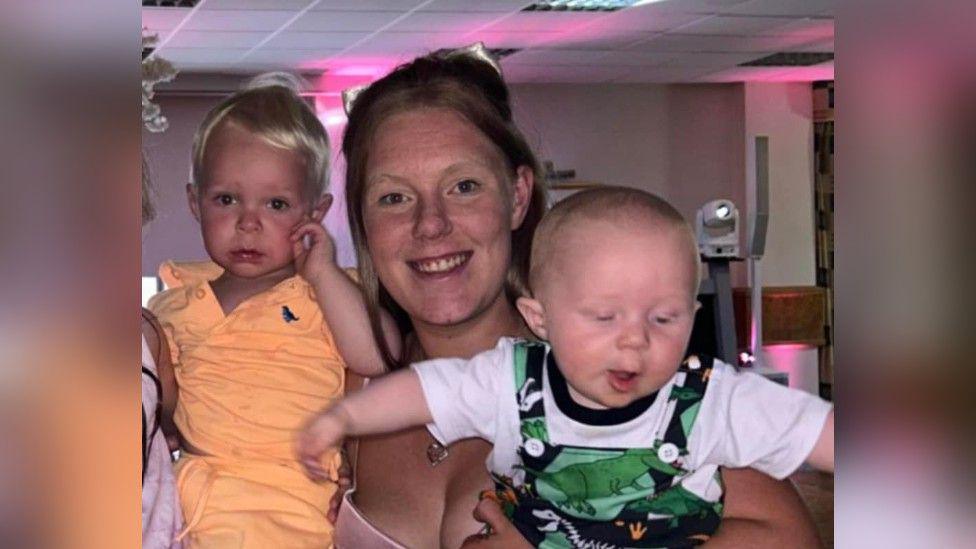 Emma Needham holds her two sons up for a photo. She is wearing a pink dress, while one son is wearing all orange, the other a white t-shirt and green overalls. 