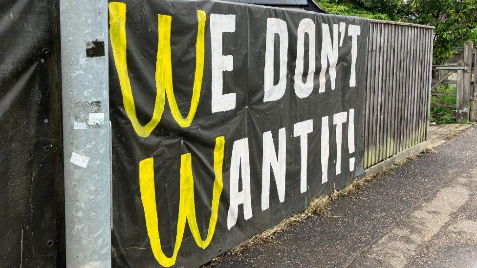 A sign attached to a fence which says WE DON'T WANT IT! in white and yellow writing.