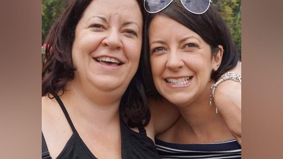 Twins with dark hair are hugging and smiling at the camera. One has sunglasses on her head and the other has a silver charm bracelet on