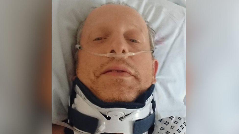 Rob Shenton takes a selfie from his hospital bed. Oxygen tubes are in his nose and he is wearing a neck brace. His grey hair appears sweaty and he looks very tired.