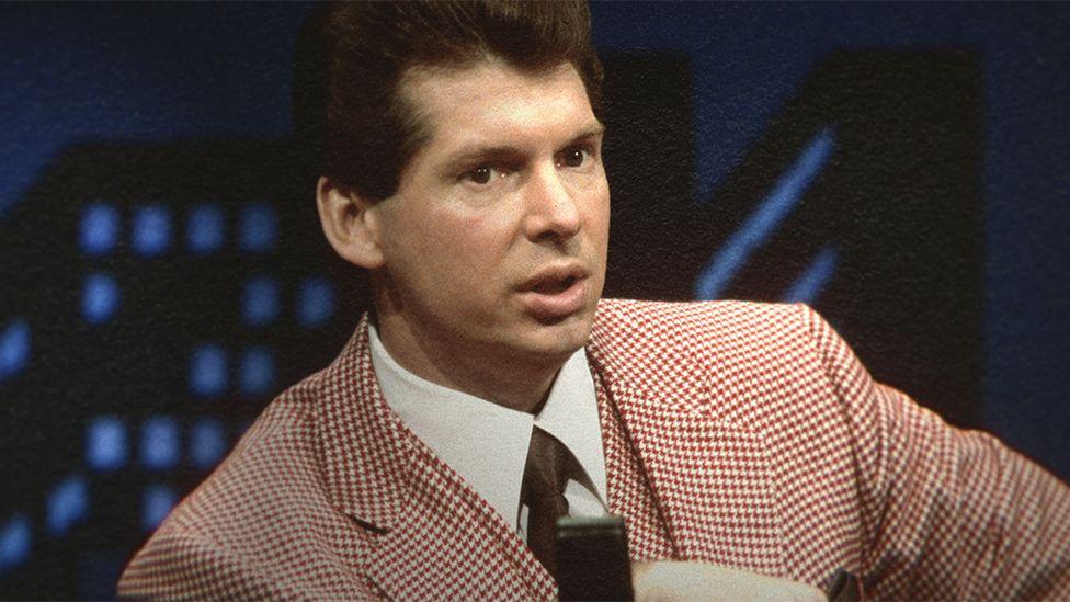 A younger Vince McMahon from the 1980s or 1990s, wearing a checked pink and white suit with white shirt and black tie.