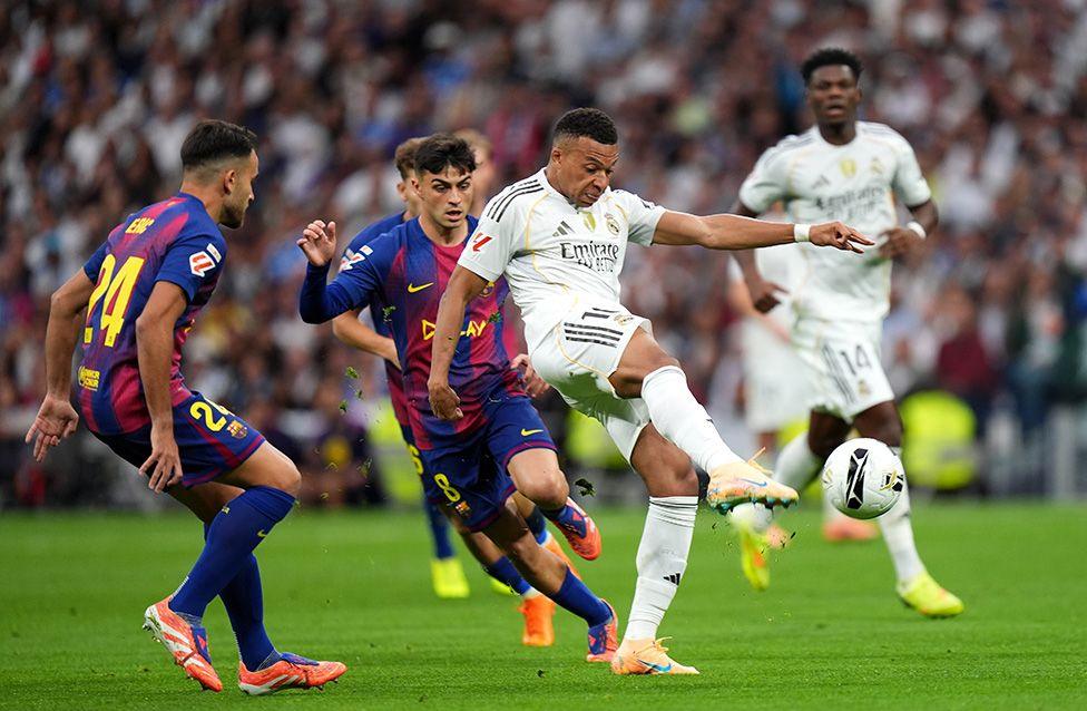 A football match shows a male player in a white Real Madrid kit kicking the ball while surrounded by three male Barcelona players in blue and red striped jerseys. The action takes place on a grass pitch with a packed stadium crowd in the background.