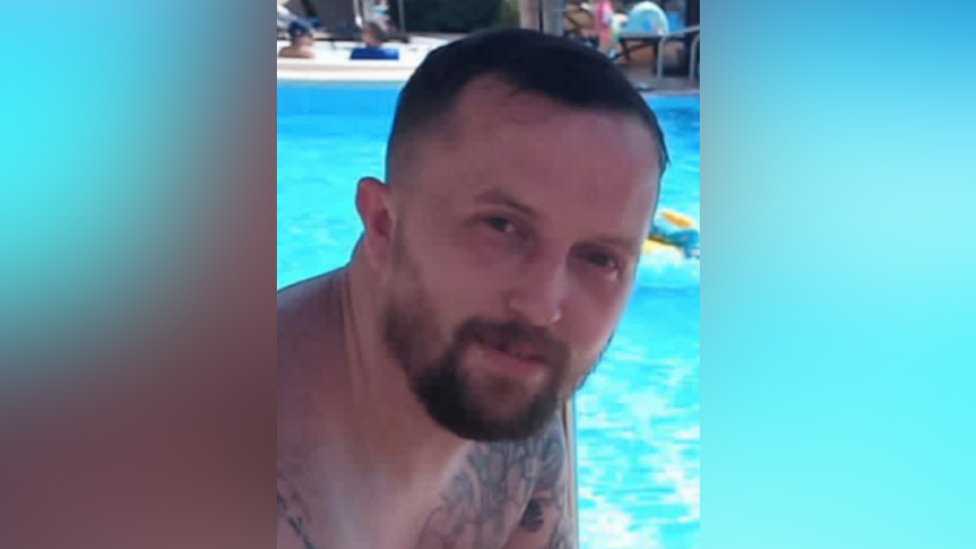 A photo of Stephen Holmes. He is shown from the shoulders up sitting in front of a pool. He has short brown hair, and a beard. He is smiling at the camera.