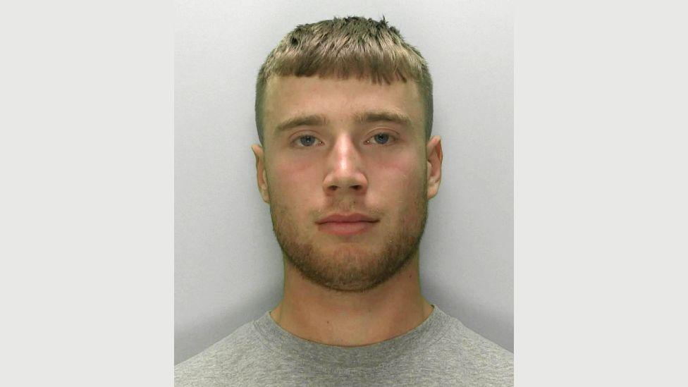 A police mugshot of Kane Pearson. He is wearing a grey T-shirt and is standing against a light grey background. He has dark blonde hair which is shaven on the sides and cut into a straight fringe on top. He has blue eyes and a dark blonde beard and is looking directly at the camera.