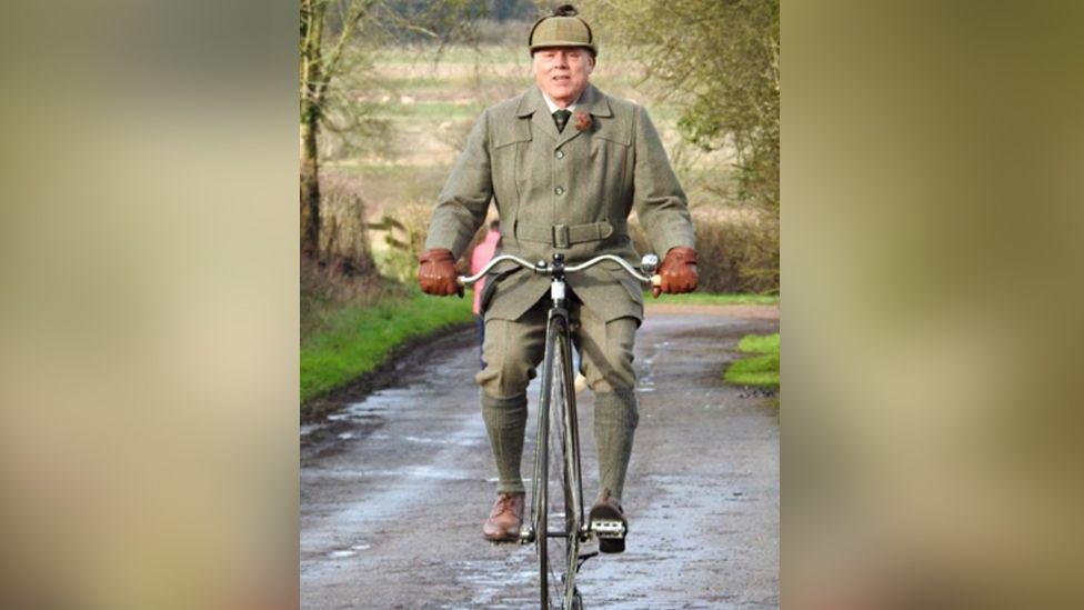 Brian Cobbold is riding his penny farthing on a country road surrounded by fields.