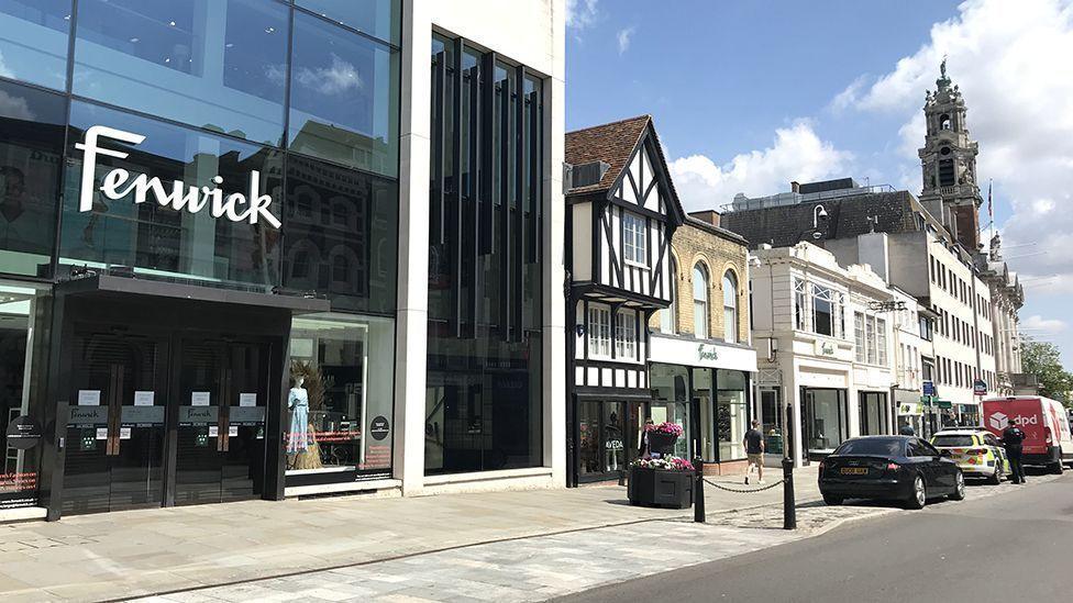 The exterior of Fenwick, as seen from Colchester High Street. The building has a mostly glass facade with the word Fenwick in front in white letters. There are cars parked in front of the building, including a police car.