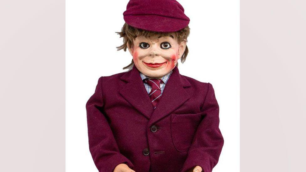 A head and torso shot of Eric Morecambe's ventriloquist dummy Charlie. It is wearing a maroon jacket over a checked shirt and a maroon tie. It has a maroon cap on its head, from which light brown hair is sticking out. It has a broad smile